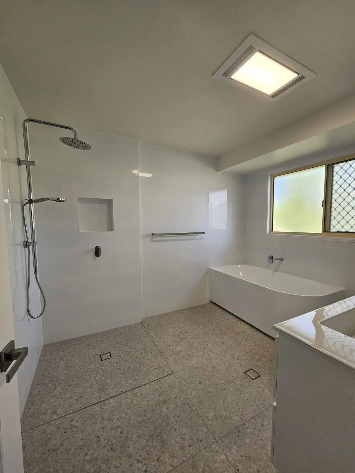 Walk-in shower with a recessed niche and rail, beside a freestanding bath, on terrazzo-look tiling