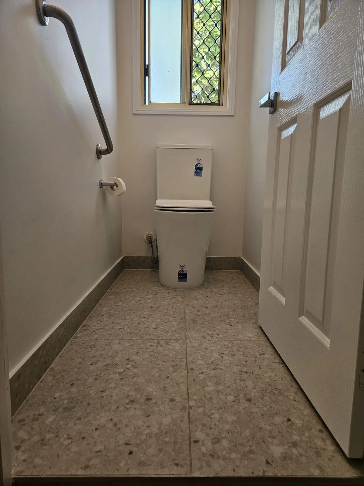 Toilet with a wall-mounted grab rail beside it, on terrazzo-look floor tiling