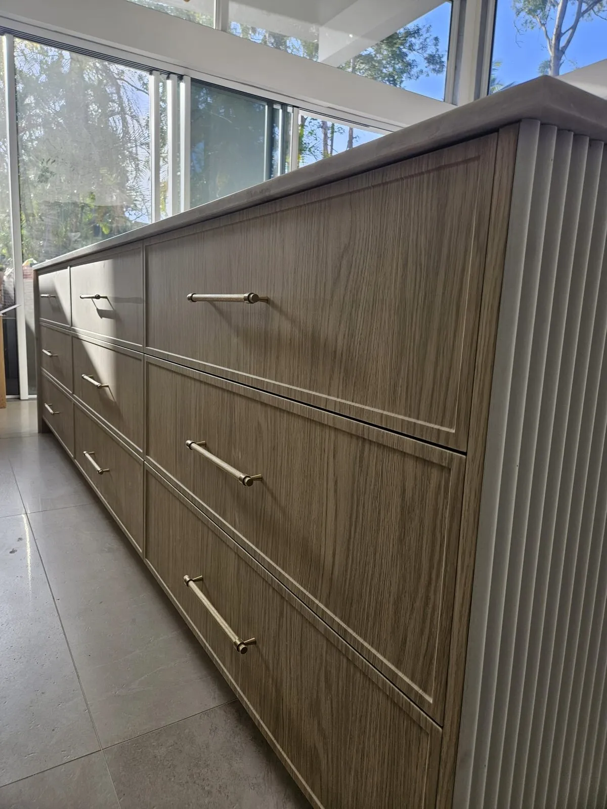 Timber drawer fronts with slim brushed-gold bar handles beside a fluted end panel, lit from a window