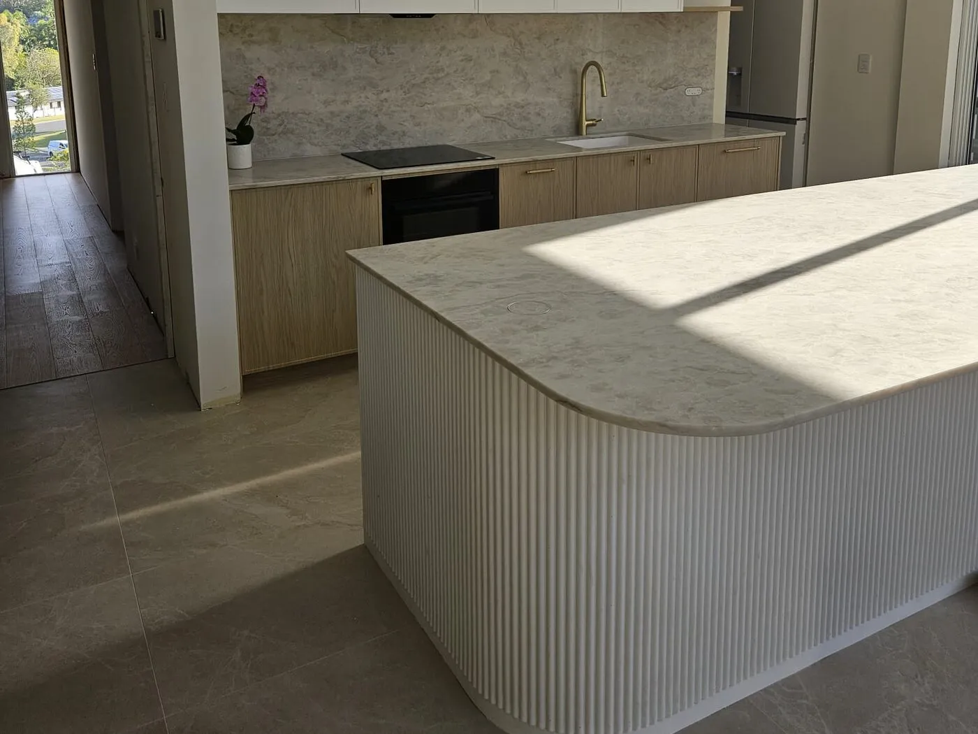 Curved fluted kitchen island with a stone benchtop, in front of a timber cabinetry run with stone splashback and brushed gold tapware