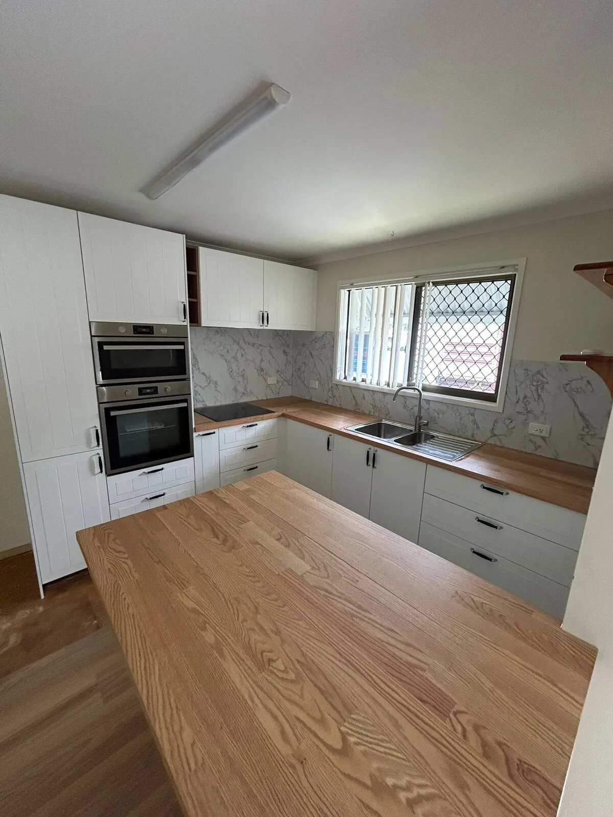 White shaker kitchen seen from the peninsula — timber benchtops on three sides, marble-look splashback, double wall oven in a full-height stack