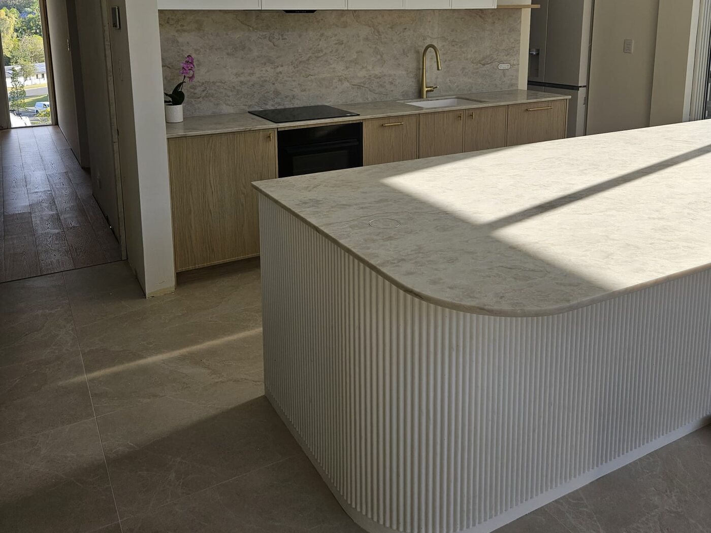 Curved fluted kitchen island with a stone benchtop, in front of a timber cabinetry run with stone splashback and brushed gold tapware