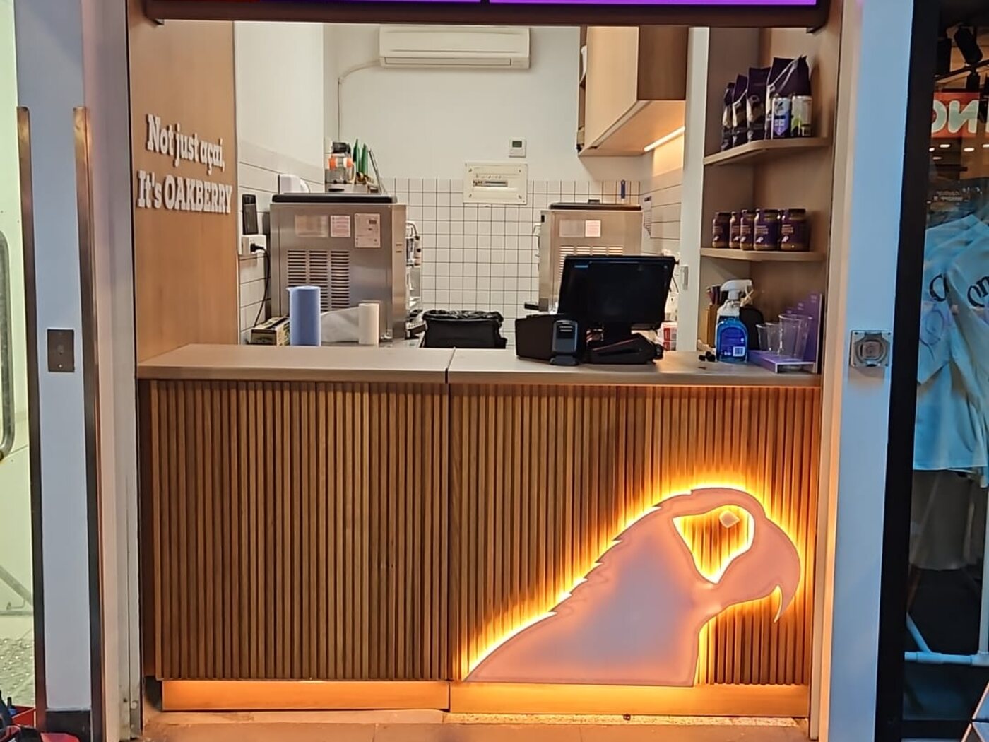 Completed Oakberry Acai kiosk fitout — timber batten servery counter with a backlit logo, shelving and menu screens