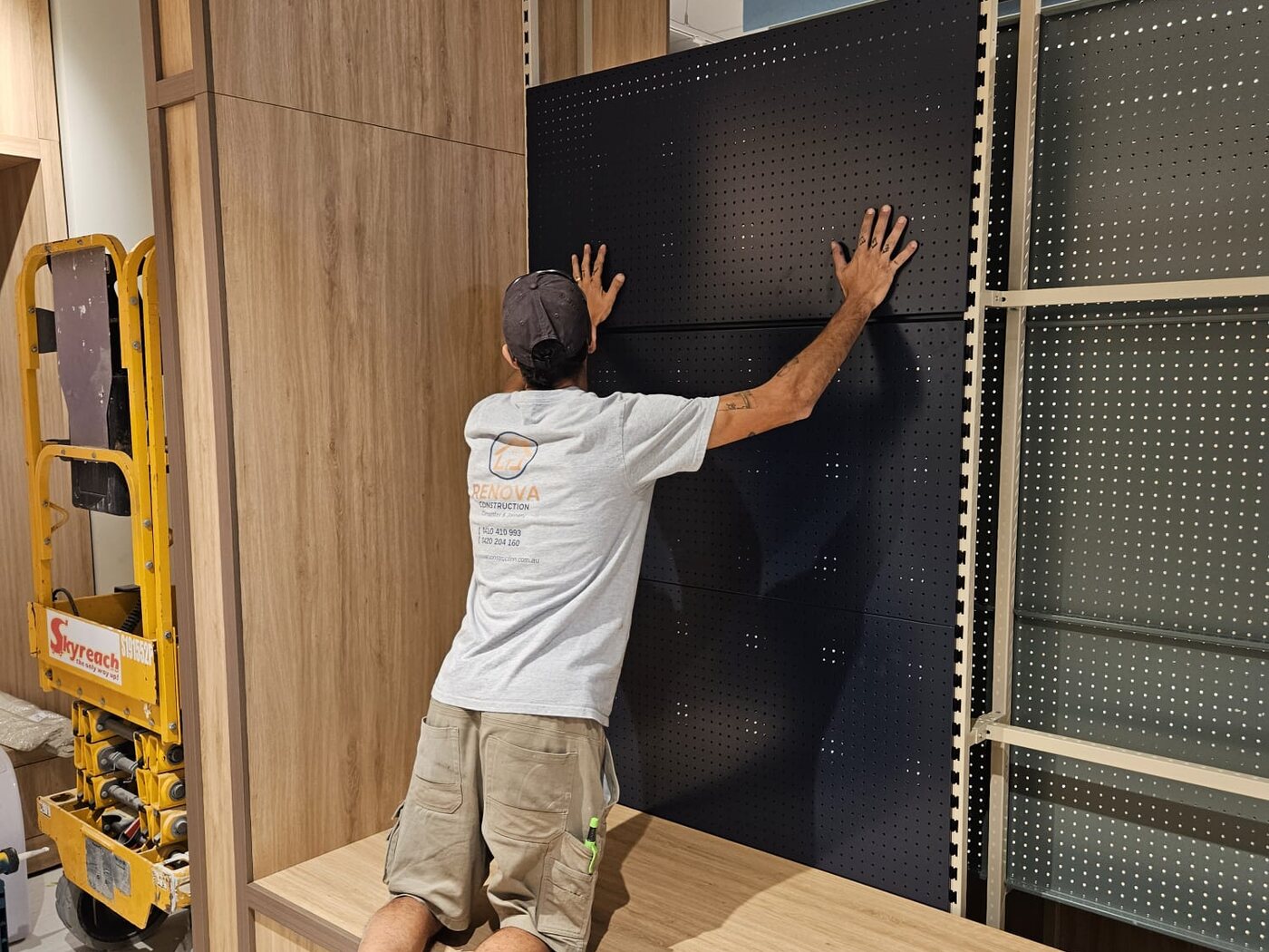 Renova carpenter fitting a perforated display panel into a retail wall system, between timber joinery, standing on the finished plinth