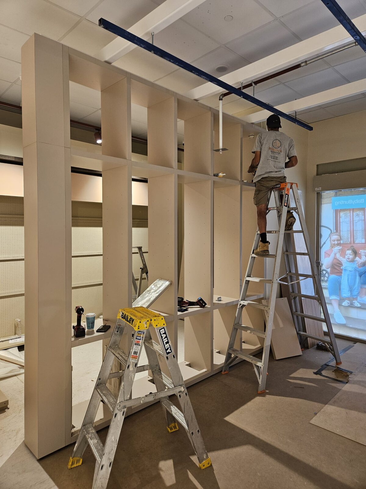 Renova carpenter on a ladder installing a full-height timber display wall in a retail fitout, ladders and tools on the floor